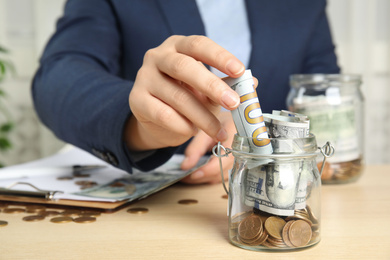 Photo of Woman putting dollar bill into jar with money on wooden table, closeup