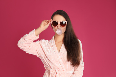 Young woman in bathrobe and sunglasses blowing chewing gum on crimson background