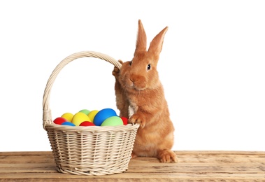 Adorable furry Easter bunny near wicker basket with dyed eggs on wooden table against white background