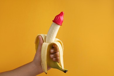 Woman holding banana in condom on orange background, closeup and space for text. Safe sex concept