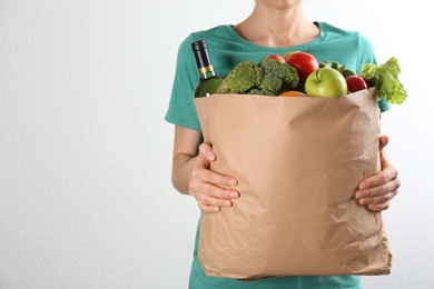 Woman holding paper bag with different groceries near white wall, closeup view. Space for text