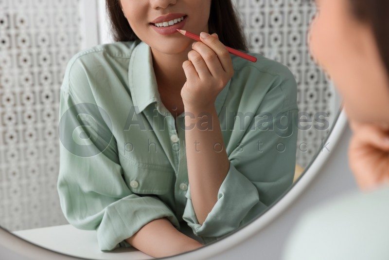 Beautiful young woman applying cosmetic pencil on lips near mirror indoors, closeup