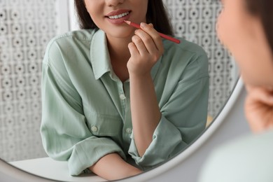 Photo of Beautiful young woman applying cosmetic pencil on lips near mirror indoors, closeup