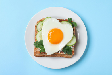 Plate of tasty sandwich with heart shaped fried egg on light blue background, top view