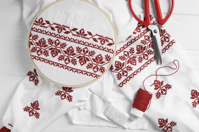 Shirt with red embroidery design in hoop, scissors and thread on white wooden table, flat lay. National Ukrainian clothes
