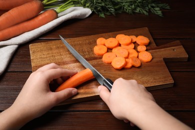 Woman cutting tasty carrot at brown wooden table, closeup