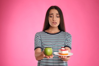 Concept of choice. Doubtful woman holding apple and cake on pink background