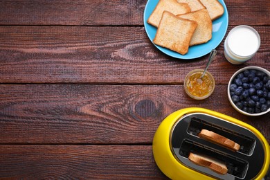 Yellow toaster with roasted bread, glass of milk, blueberries and jam on wooden table, flat lay. Space for text
