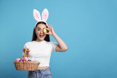 Photo of Happy woman in bunny ears headband holding wicker basket of painted Easter eggs on turquoise background. Space for text