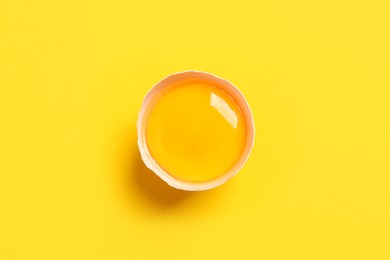 Cracked eggshell with raw yolk on yellow background, top view