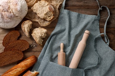 Clean kitchen apron with rolling pins and different types of bread on wooden table, flat lay