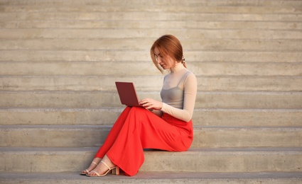 Beautiful young woman with laptop sitting on stairs outdoors