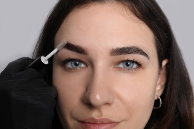Beautician fixing woman's eyebrows with gel after tinting on light grey background, closeup