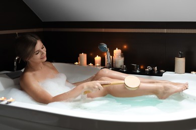 Beautiful woman rubbing leg with brush while taking bubble bath. Romantic atmosphere