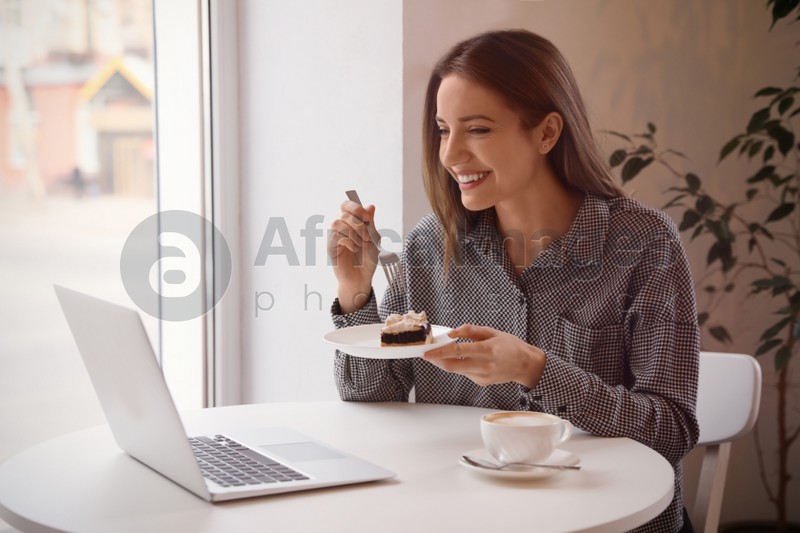 Young blogger with laptop eating dessert at table in cafe