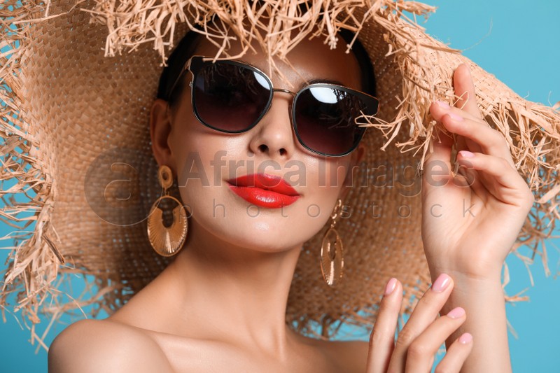 Attractive woman in fashionable sunglasses and wicker hat against light blue background, closeup
