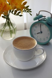 Photo of Morning coffee, alarm clock and flowers on white table indoors