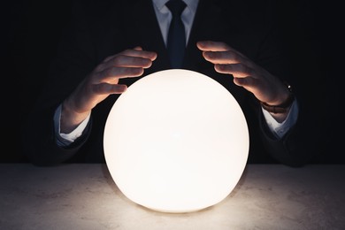 Businessman using glowing crystal ball to predict future at table in darkness, closeup. Fortune telling