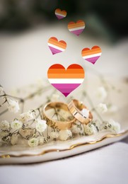 Illustration of hearts in color of lesbian flag, beautiful wedding rings and gypsophila flowers on white cloth, closeup view