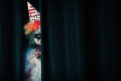 Terrifying clown hiding behind black curtains, space for text. Halloween party costume