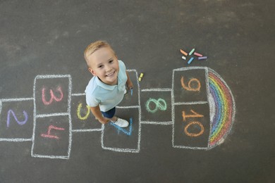 Little boy and colorful hopscotch drawn with chalk on asphalt outdoors, top view. Happy childhood