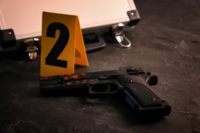 Gun with blood and evidence marker on black slate table, closeup. Crime scene