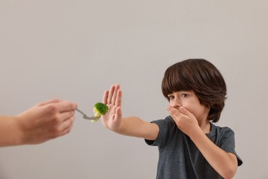 Cute little boy covering mouth and refusing to eat broccoli on grey background