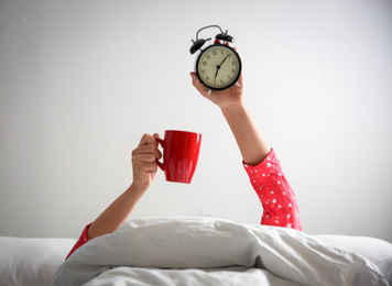 Woman with cup and alarm clock lying in bed, closeup. Morning time