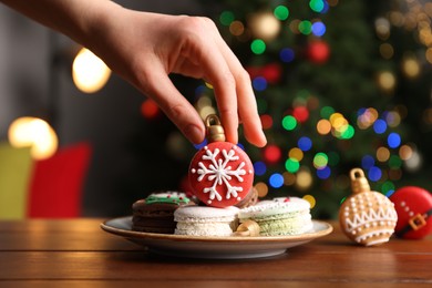 Photo of Woman taking Christmas macaron from plate at wooden table against blurred festive lights, closeup