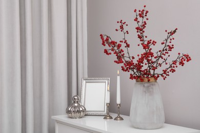 Hawthorn branches with red berries in vase, candles and frame on table indoors, space for text