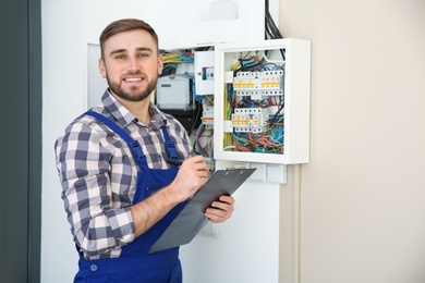 Male electrician with clipboard near fuse board indoors, space for text
