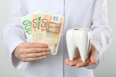 Dentist holding ceramic model of tooth and euro banknotes on light background, closeup. Expensive treatment
