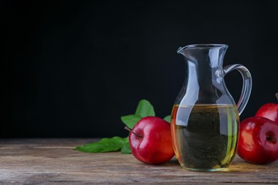 Jug of tasty juice and fresh red apples on wooden table against black background, space for text