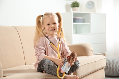 Cute child imagining herself as doctor while playing with stethoscope and doll on couch at home