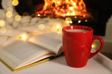 Cup of hot drink and book near fireplace. Cozy Christmas atmosphere