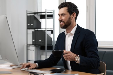 Handsome bearded man with cup of drink working on computer at table in office