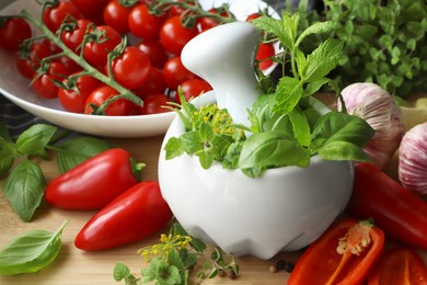 Mortar with fresh herbs near garlic, pepper and cherry tomatoes on wooden table, closeup