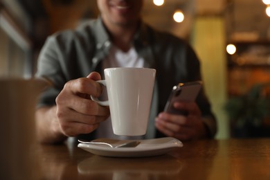 Man with cup of coffee and smartphone at table in morning, closeup