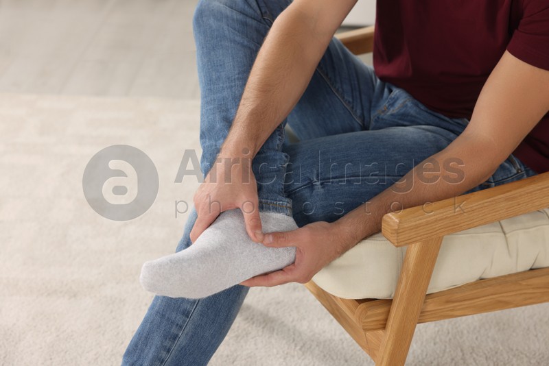 Photo of Man massaging leg on soft armchair indoors, closeup