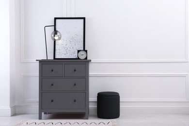Grey wooden chest of drawers with lamp, clock and picture near white wall in room, space for text. Interior design