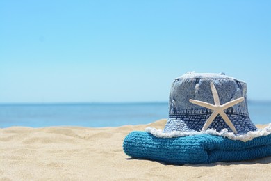 Stylish denim hat, towel and starfish on sand near sea, space for text. Beach accessories