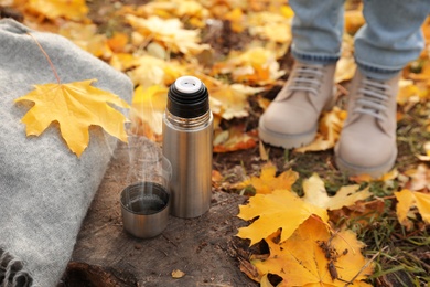Woman standing near metallic thermos, cap and scarf on tree stump in autumn park, closeup