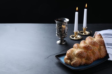 Photo of Homemade braided bread with sesame seeds, goblet and candles on dark grey table, space for text. Traditional Shabbat challah