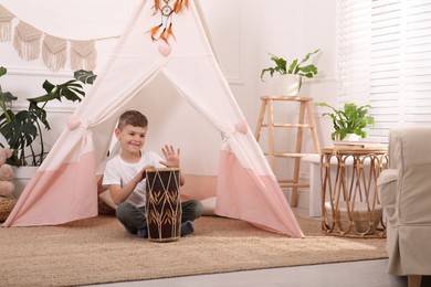 Cute little boy playing drum near toy wigwam at home