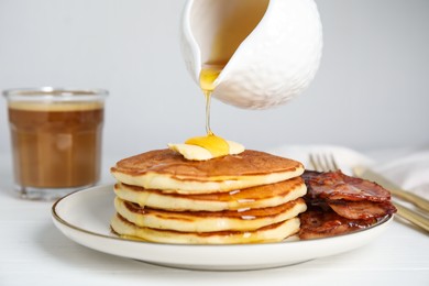 Pouring maple syrup onto delicious pancakes with butter and fried bacon at white wooden table, closeup