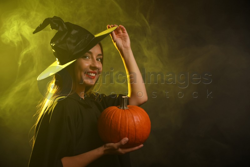 Young woman wearing witch costume with pumpkin in smoke cloud on dark background, space for text. Halloween party