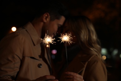 Photo of Couple in warm clothes holding burning sparklers at night, focus on fireworks