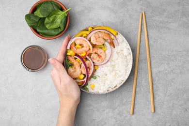 Photo of Woman holding delicious poke bowl with shrimps, rice and vegetables at light grey table, top view