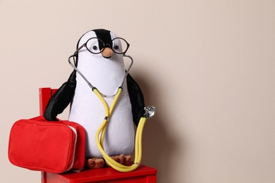 Toy penguin with eyeglasses, stethoscope and first aid bag on beige background, space for text. Pediatrician practice