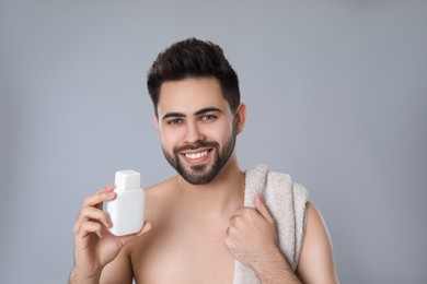 Handsome young man with beard holding post shave lotion on grey background
